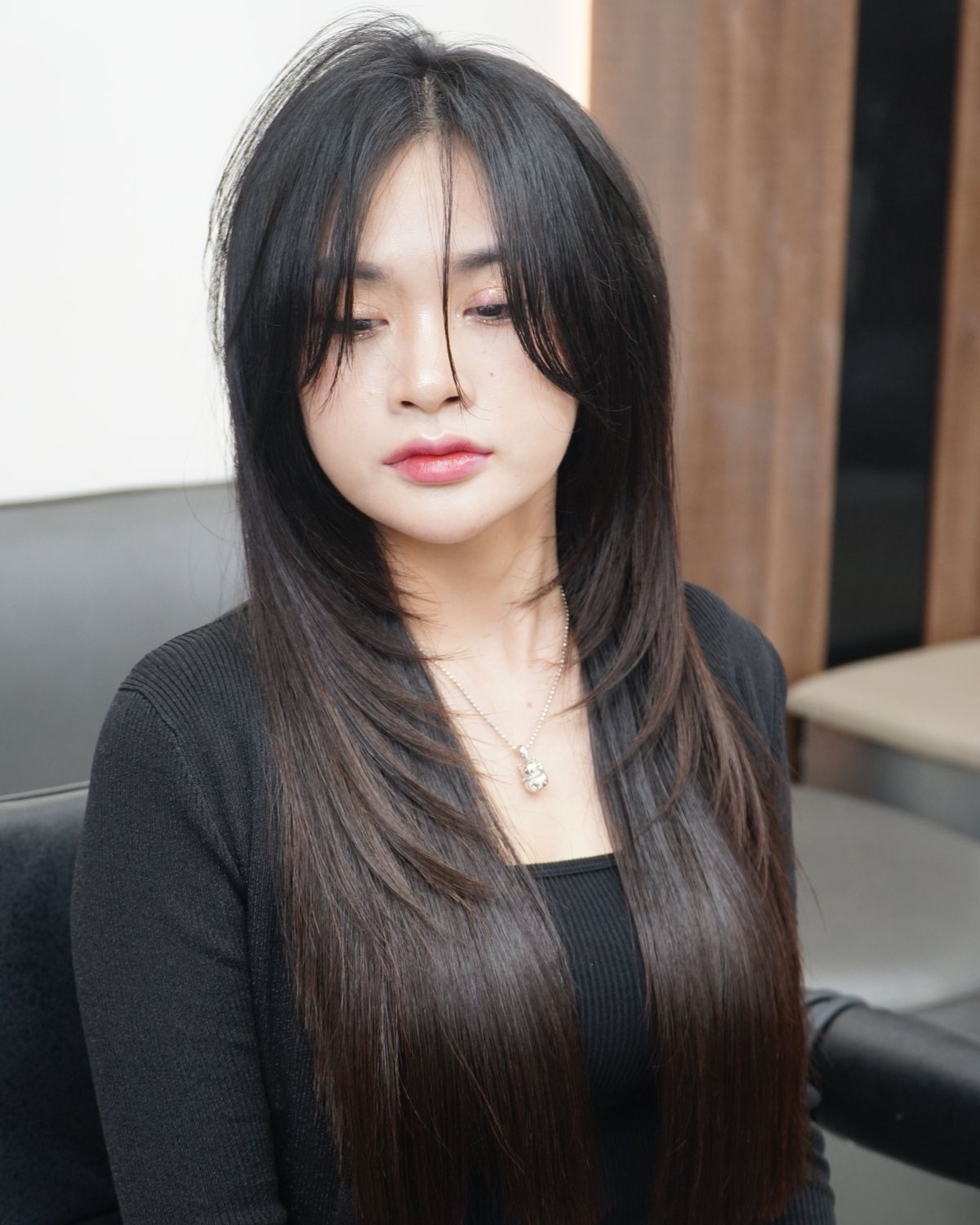 Woman with long, sleek black layered hair and a black cardigan, sitting in a softly lit salon.