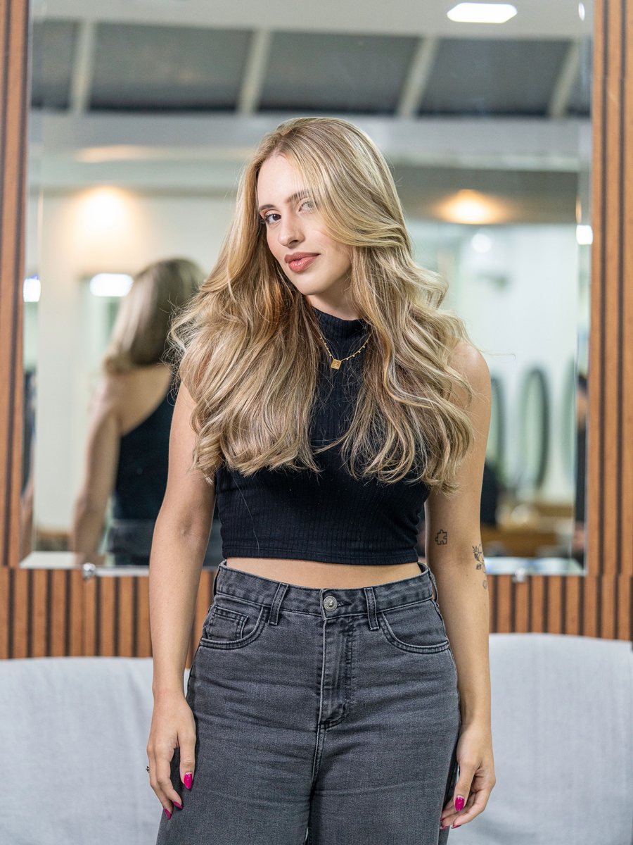 Smiling stylist with long blonde waves and a black top, in front of a salon mirror.