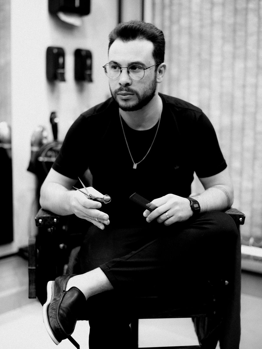 Hairstylist in a black T-shirt and glasses, seated with scissors and a comb, in black and white.