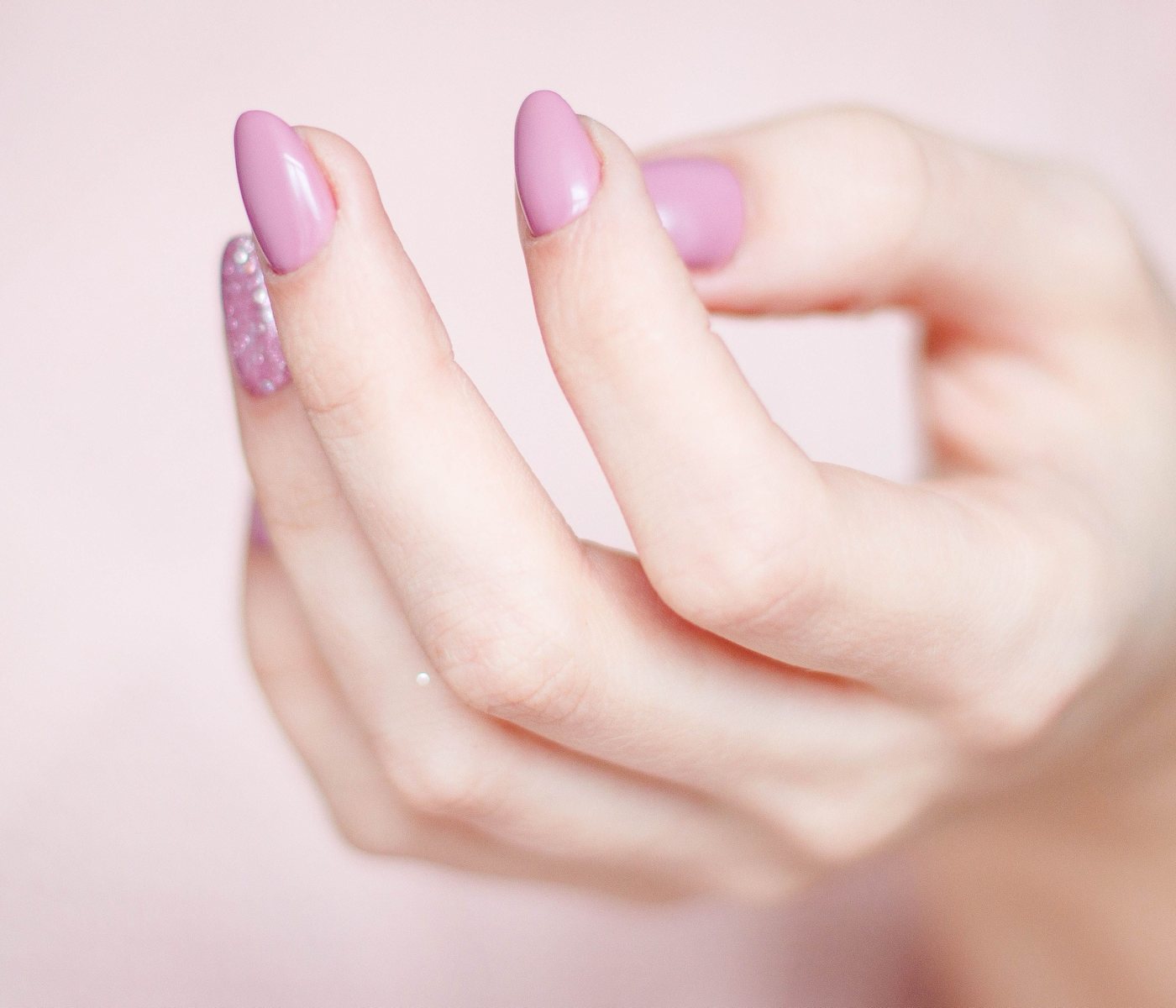 A hand with glossy pink almond nails and one glitter accent nail, against a soft pink background.
